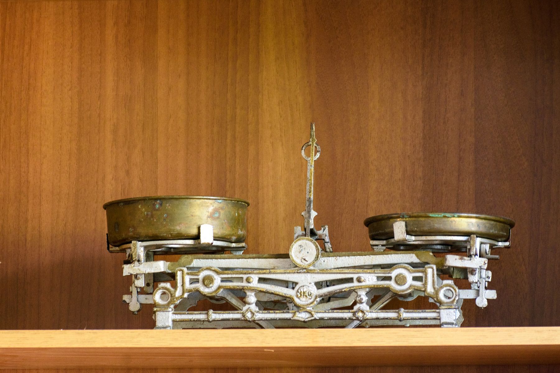 An old two-pan balance scale, both brass pans empty, standing level on a wooden shelf.