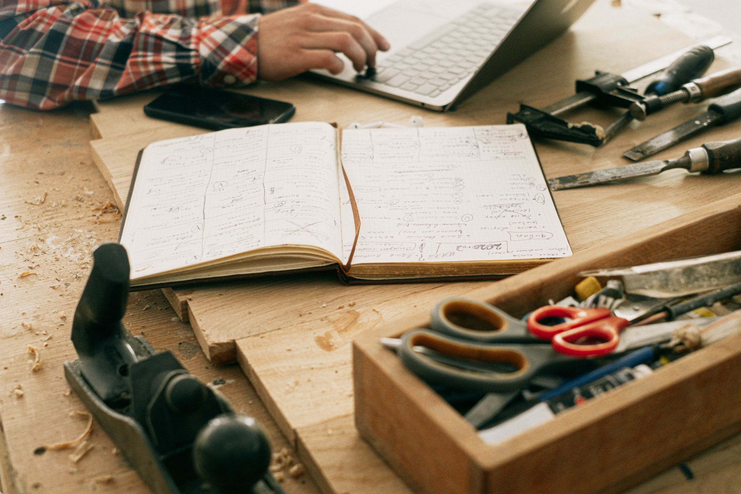 A workbench of pale wood covered in shavings: an open notebook filled with handwriting, a laptop and a hand at the keyboard behind it, a hand plane, and a tray of chisels and scissors.
