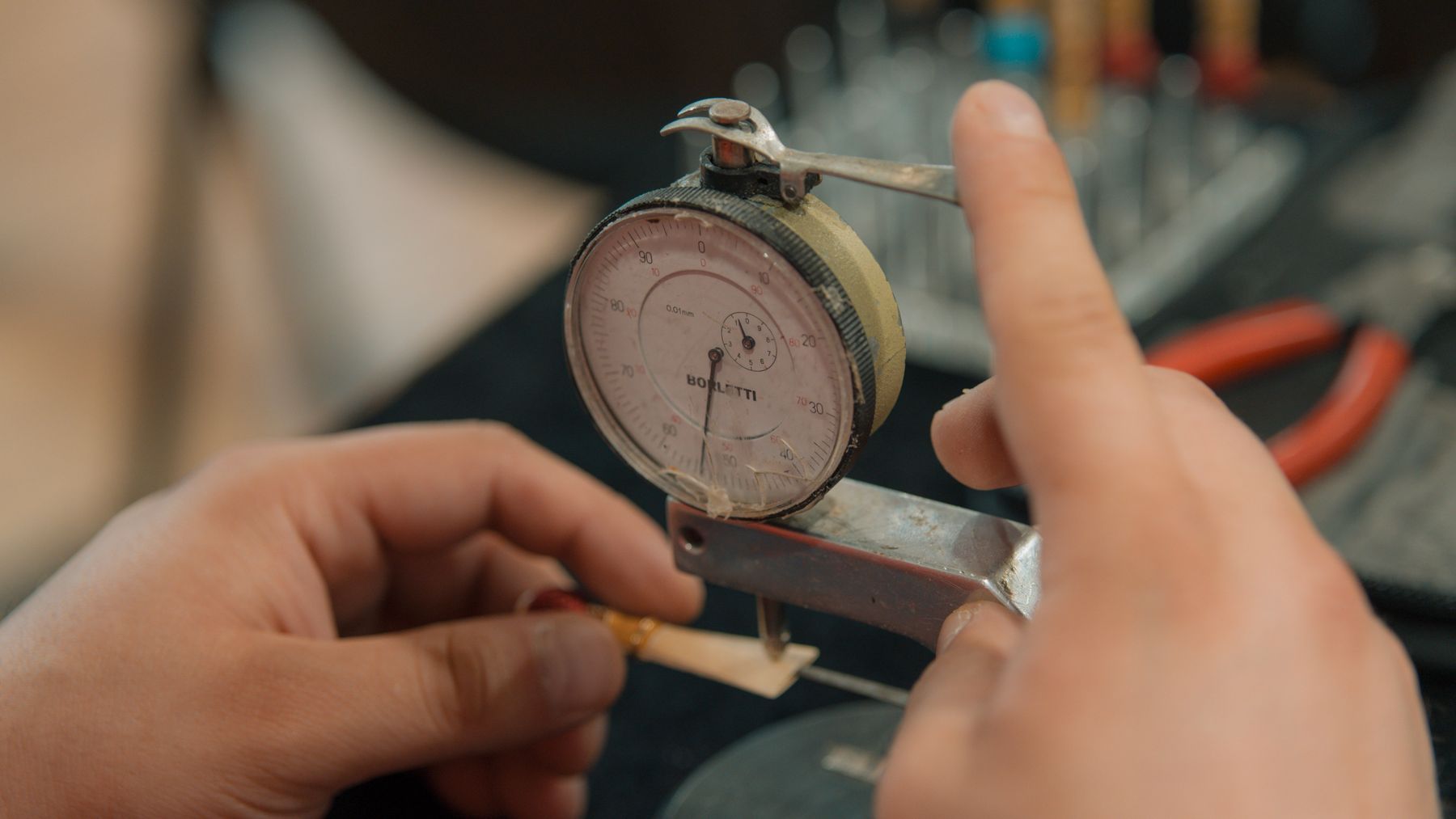 Two hands holding a dial indicator against a piece of metal, the needle resting mid-scale, workshop tools out of focus behind.
