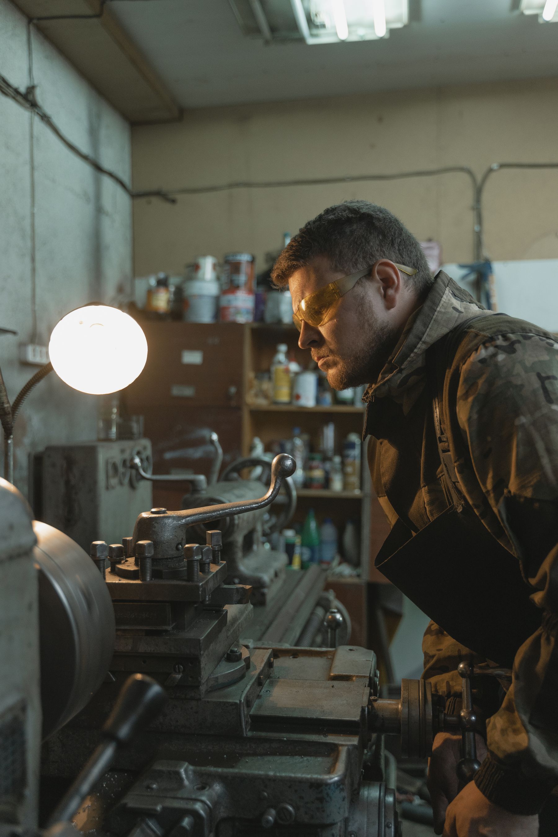 A machinist in safety glasses leaning over a metal lathe under a work lamp, watching the cut, shelves of tools and bottles behind him.