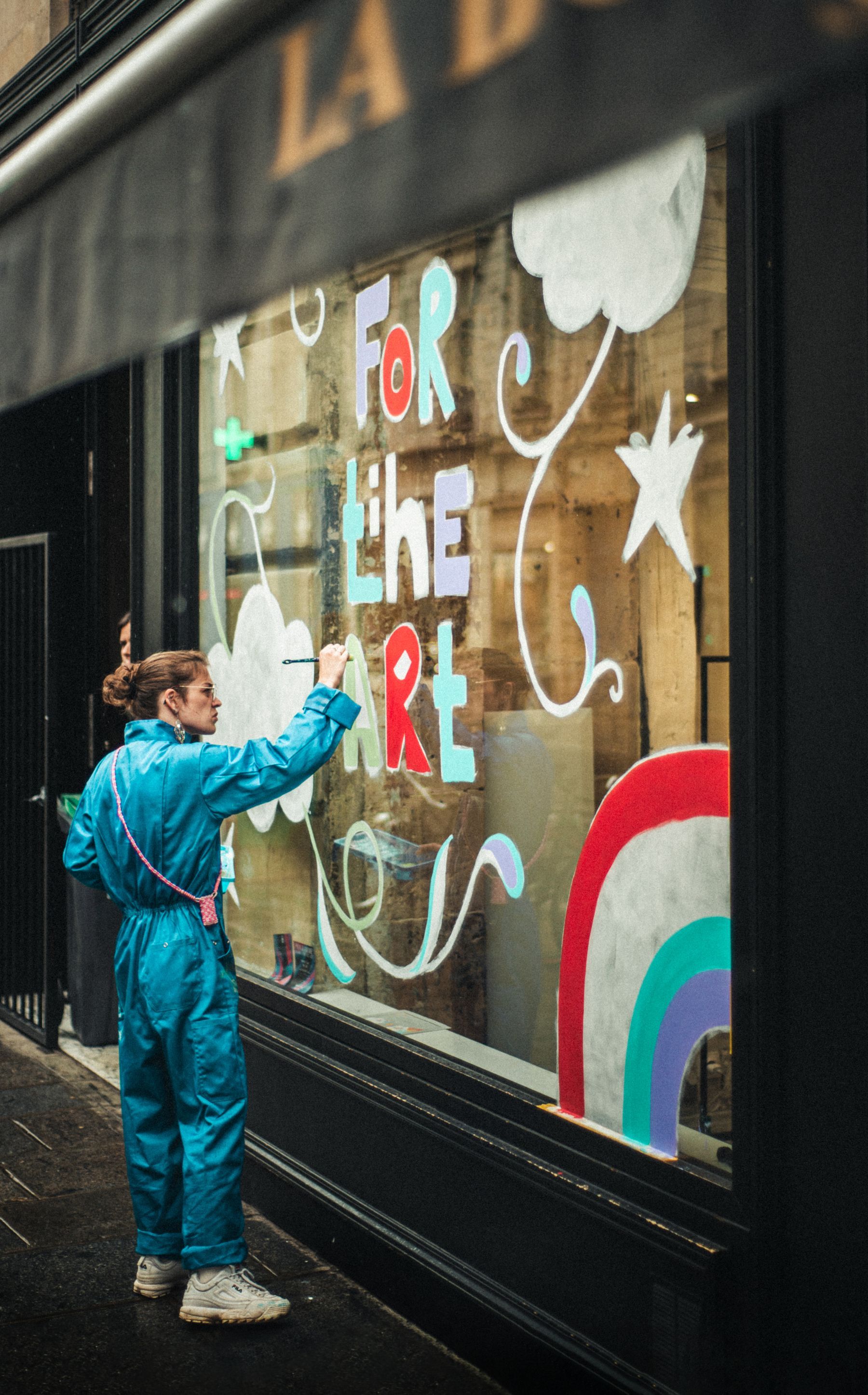 A signwriter in a blue boilersuit, brush raised, painting bold hand-lettering and a rainbow onto a shop window from the pavement outside.