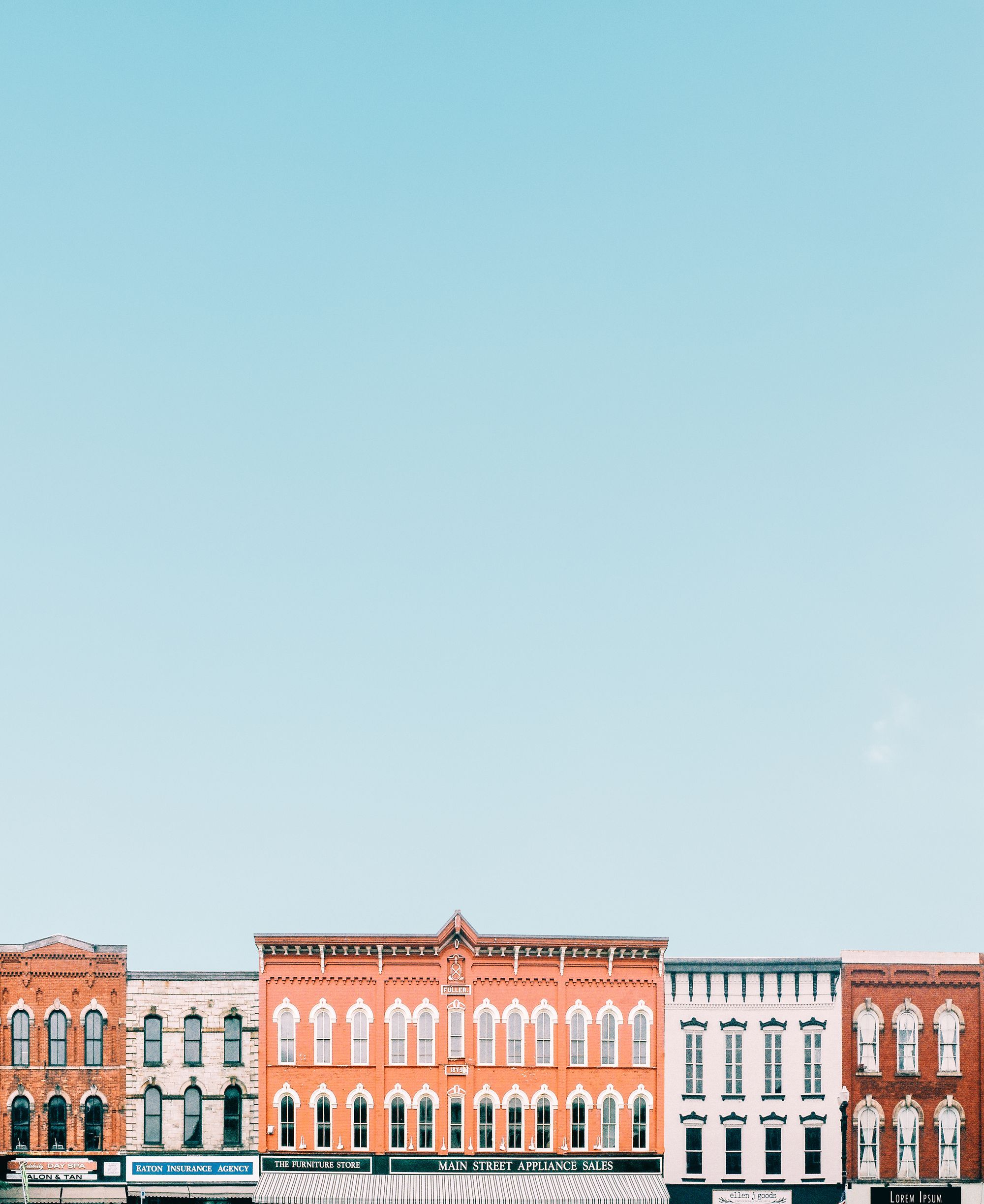 A row of two-storey main-street buildings under a wide pale sky, their ground floors lettered with the names of independent businesses: an insurance agency, a furniture store, an appliance dealer.